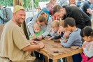 Ein Handwerker in traditioneller Kleidung bietet an einem Tisch ein Bastelangebot für Kinder an. Kinder und Erwachsene sitzen und stehen um ihn herum und werkeln. 