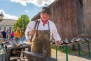 Ein Mann in Lederschürze und Hut hält einen Hammer in der Hand, mit dem er bereit ist auf ein Eisenstab in sei-ner anderen Hand zu schlagen. Hinter ihm auf einem Tisch sind verschiedene gusseiserne Pfannen platziert.