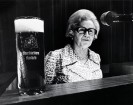 Ein schwarz-weiß Foto von einer Frau mit kurzen, frisierten Haaren und eine dunklen Brille, die in ein Mikrofon spricht. Vor ihr abgestellt steht ein Glas Kölsch.