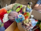 Kleine Püppchen gebastelt aus Draht, die platziert sind auf Kartonkisten, in denen sich Bastelmaterial und Woll-knäuel befinden. 