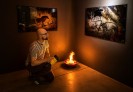 In einem kleinen Raum brennt eine Flamme als Lichtquelle. An der Wand hängen zwei Bilder. Ein Mann in einem gestreiften T-Shirt kniet davor und hält eine große, gelbe Taschenlampe auf sie. 