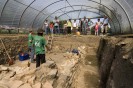 Eine Gruppe Besucher*innen steht vor einer Grabungsgrube, in der zwei Archäolog*innen stehen und ihre Arbeit vorstellen.