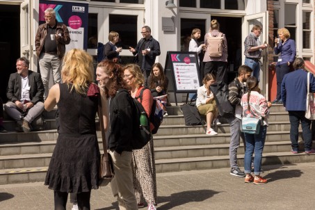 Menschen stehen und sitzen vor einem Gebäude mit offenen Türen auf Treppen oder stehen in Gruppen zusammen. Es sind Aufsteller der LVR-Kulturkonferenz 2025 zu sehen.