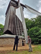 Zwei Personen stehen vor einer großen Holzmühle. Um sie herum stehen Bäume. 