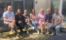 Eine Gruppe aus sieben Personen sitzt auf zwei Metallbänken vor einem Haus.