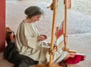 Eine Frau mit Haarnetz und weißem, mittelalterlichem Gewand sitzt vor einem Webstuhl. Sie schaut auf ihre Hände,mit denen sie ihr Handwerk ausführt und lächelt. 
