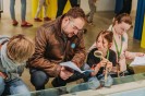 Zwei Erwachsene und zwei Kinder knien vor einer Museumsvitrine und lesen in einem schmalen Heft. Die Kinder haben Lupen, Stifte und Lineale bei sich.