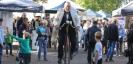 Ein Mann im Anzug und weißem Mantel fährt ein historisches Fahrrad an verschiedenen Ständen vorbei. Einige Menschen schauen ihm dabei zu.