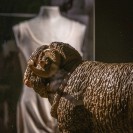 Eine Figur von einem Schaf. Dahinter schemenhaft ein weißes, dünnes Kleid auf einer Ausstellungspuppe ohne Kopf. 