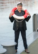 Ein Mann steht an einer Reling. Er trägt einen dunklen Anzug, eine Kapitänsmütze auf dem Kopf und einen rotweißen Schwimmring um die Schultern.