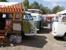 Bunte VW-Busse auf einem Campingplatz. Einige haben die Seitentüren geöffnet und Gegenstände, wie Stühle oder einen Koffer, davorgestellt.