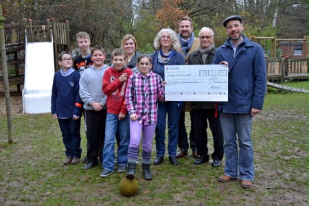 Gruppe von Erwachsenen und Kindern auf Spielplatz. Erwachsene halten einen übergroßen Scheck in der Hand.