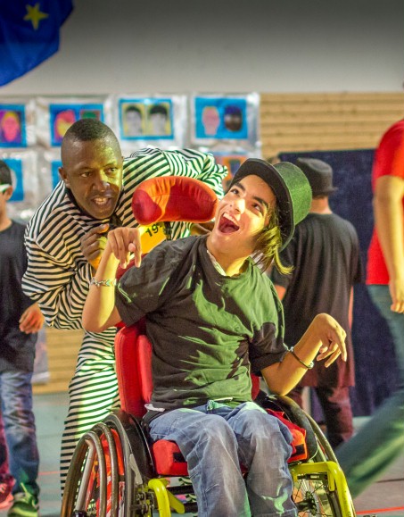 Ein Mann steht hinter einem Jungen, der in einem Rollstuhl sitzt. Beide lachen in die Kamera.