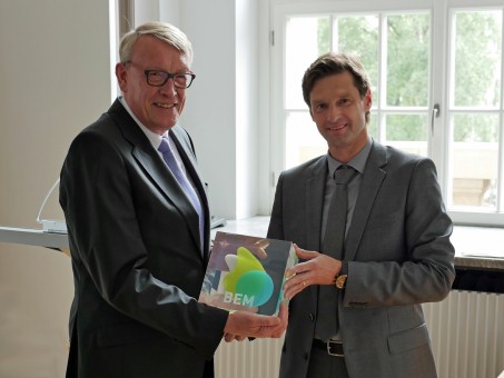 Zwei Männer halten einen Acrylwürfel in der Hand und schauen in die Kamera.