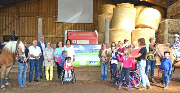 Gruppe von Erwachsenen, Tieren und Kindern in einem Reitstall vor einem Banner