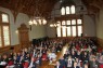 Viele Menschen sitzen in einem großen, historischen Saal mit hoher Decke und hören einem Vortragenden zu.