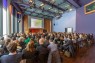 Ein Saal mit vielen sitzenden Menschen, die auf eine Bühne blicken.