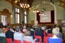 Viele Personen sitzen in einem Saal, von hinten fotografiert und schauen sich eine Präsentation an.
