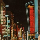 Ansicht einer Einkaufstraße der 60er Jahre bei Nacht, mit hell erleuchteten Reklametafeln der Geschäfte.