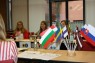 Europäische Flaggen stehen in einer Halterung auf einem Tisch. An den Tischen sitzen mehrere Personen im Hintergrund.