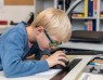 Ein Junge mit Brille sitzt an einem Schreibtisch und benutzt ein Vergrößerungsglas zum Lesen.