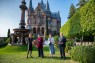 Drei Frauen und ein Mann auf einer Wiese vor einem Schloss. 