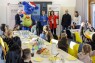 Man sieht eine Gruppe von Kindern und Eltern an einer langen Tafel. Davor stehen sechs Personen vor zwei Roll-Ups des LVR. 