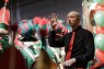 Man sieht einen Mann mit Krawatte, der Gebärdensprache spricht. Dahinter stehen mehrere Personen in grün-roter Uniform. 