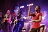 Man sieht eine Frau, die Gebärdensprache spricht. Dahinter spielt eine Band Musik. 