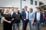 Man sieht eine Gruppe von Menschen, die vor einem Gebäude stehen. Daneben sieht man weitere Menschen auf einer Eröffnungsfeier.