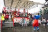 Blick vom Zugweg auf eine große Tribüne, auf der zahlreiche Menschen in Kostümen gekleidet die Arme hochhalten und dem Maskottchen Mitmän zuwinken.