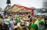 Man sieht eine Tribüne mit vielen verkleideten Menschen. Davor geht der Rosenmontagszug entlang. 