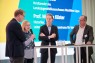 Drei Männer und eine Frau auf einem Podium. Die Frau spricht in ein Mikrofon