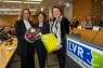 Man sieht drei lächelnde Frauen in einem Saal voller Menschen. Die beiden Frauen am Rand halten einen Blumenstrauß und ein verpacktes Geschenk in die Höhe. 