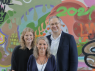 Zwei blonde Frauen und ein Mann mit Brille vor einer Wand mit Graffitti