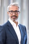 Ein Mann mit grauen Haaren, Brille und Anzug lächelt in die Kamera.