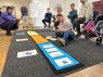 Eine Frau mit Down-Syndrom sitzt auf dem Boden und ordnet Karten einem Zeitstrahl auf dem Boden zu. Ein Tontechniker hockt daneben und hält ein Mikrofon.