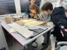 Zwei junge Frauen beugen sich über einen von unten angeleuchteten Tisch, auf dem alte Papierdokumente liegen.