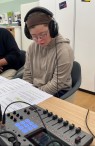 Eine Frau mit Down-Syndrom trägt Kopfhörer und spricht einen Text in ein Mikrofon. Im Vordergrund ein Aufnahmegerät.