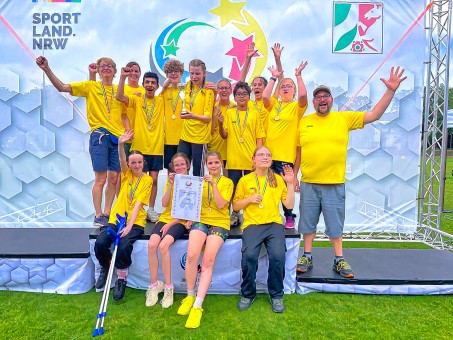 Ein Gruppe Schülerinnen und Schüler mit gelben Trikots lächeln und winken in die Kamera.
