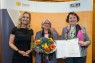 Drei Frauen stehen vor einer orangenen Fotowand und lächeln in die Kamera.