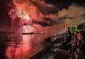 Zu sehen sind Menschen, die in der Dunkheit am Rhein sitzen und sich über dem Wasser ein Feuerwerk anschauen. Auf dem Wasser sind Schiffe zu sehen.