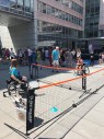 Zu sehen sind ein Junge und ein Mädchen im Rollstuhl, die miteinander Rollstuhl-Tennis spielen. Im Hintergrund sind Menschen zu sehen.