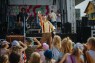 Zu sehen ist ein verkleideter pantomimischer Künstler mit einer Glaskugel in der Hand. Viele Kinder, die von hinten zu sehen sind, schauen seiner Show zu. Im Hintergrund ist eine Bühne zu sehen.