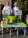 Zwei Frauen stehen nebeneinander und l&auml;cheln in die Kamera. Sie halten jeweils eine Tafel hoch mit dem Logo und Informationen zur Veranstaltung. Vor ihnen steht eine Bank mit gr&uuml;nen Kissen, auf der ein Hund liegt.