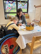 Ein Kind sitzt in einem Rollstuhl und malt mit einer Frau zusammen an einem Tisch