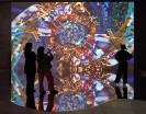 Drei Menschen stehen vor einer bunten Videoinstallation. Der Boden auf dem sie stehen ist mit Spiegelfolie beklebt, um den Effekt der Projektion zu verstärken.