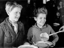 Eine schwarz-weiße Fotografie auf der zwei lächelnde Kinder zu sehen sind, die Teller in den Händen halten und gerade etwas zu essen bekommen.