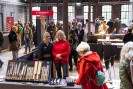 Teilnehmer*innen und Austeller*innen in einer Halle in der verschiedene Stände Produkte präsentieren, unter anderem Messer.