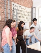 Besucher*innen in einer Ausstellung. Hinter ihnen befindet sich ein Zaun und Plakate mit Text „Kein Mensch bekämpft die Freiheit, er bekämpft höchstens die Freiheit der anderen (Karl Marx).“